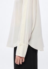 Fehér ing hosszú ujjal, gombos mandzsettával és oldalsó hasítékokkal. Puha, könnyű anyag sima textúrával. Egyszerű dizájn.