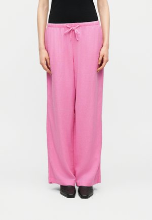 Personne portant un pantalon rose ample à cordon, un haut noir et des chaussures noires pointues, debout devant un fond clair uni.