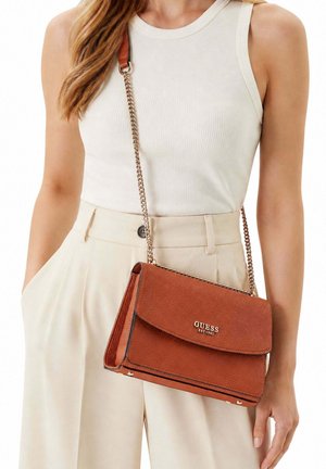 Femme portant un top blanc sans manches et un pantalon beige, avec un sac à bandoulière Guess en cuir marron à chaîne dorée.