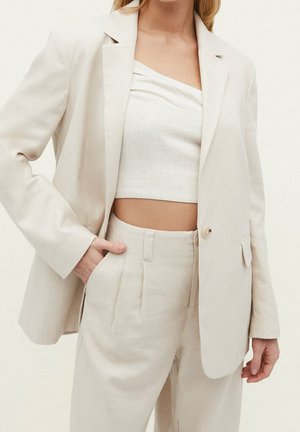 Femme portant un blazer beige clair sur un top court assorti et un pantalon taille haute, une main dans la poche, sur un fond uni.