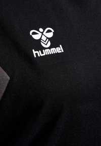 Sort stof t-shirt med et hvidt Hummel-logo. Designet inkluderer et bi-emblem og geometriske detaljer på skjortens stof.