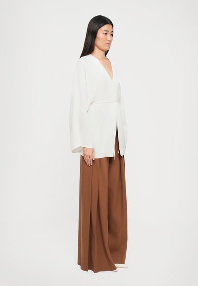 Woman with long dark hair wearing a loose white V-neck top with wide sleeves and brown wide-leg pants, standing against a white background.
