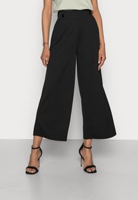 Pantalons larges noirs à taille haute, avec deux boutons, fabriqués dans un tissu lisse. Associés à des sandales noires à talons aiguilles à lanières.
