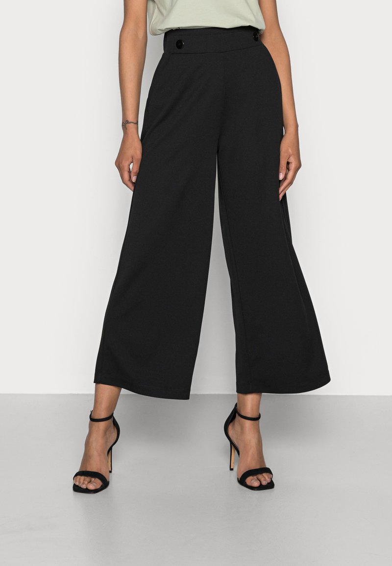 Pantalons larges noirs à taille haute, avec deux boutons, fabriqués dans un tissu lisse. Associés à des sandales noires à talons aiguilles à lanières.