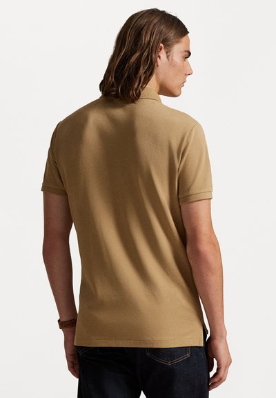 Μπεζ πόλο μπλουζάκι με υφή, κοντά μανίκια και πλαϊνές σχισμές, σχεδιασμένο για άνετη εφαρμογή. Η πίσω όψη δείχνει μια λεία υφή και διακριτή ραφή.