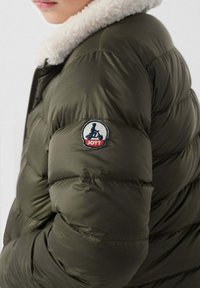 Giubbotto puffer verde oliva con un morbido colletto in shearling bianco, caratterizzato da un'etichetta circolare con il logo JOTT sulla manica sinistra. Tessuto testurizzato.