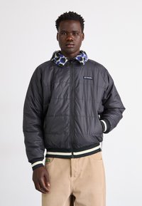 Schwarze Steppjacke mit einem gemusterten Fleece-Kragen, Reißverschluss vorne und gestreiften Bündchen. Verfügt über ein Logo-Patch auf der Brust und ein gesteppter Design.