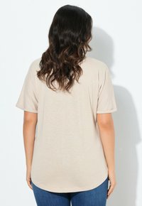 Camiseta de manga corta beige con un ajuste relajado, presenta un dobladillo redondo y un material ligero y texturizado. La vista trasera muestra sutiles variaciones en la tela.