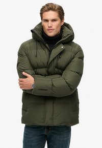 Oliven grøn pufferjakke med hætte, quiltet design, justerbare snore og sidelommer. Båret over en sort turtleneck sweater.