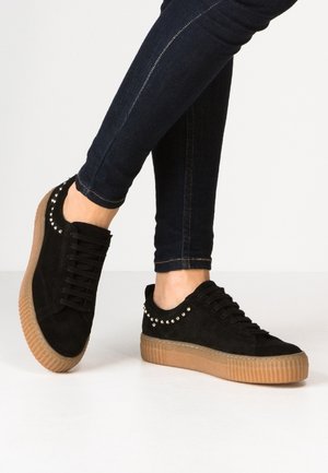 Sorte ruskindssneakers med brune gummi såler og sølvnitter nær hælen, båret med mørke skinny jeans på krydsede ben.