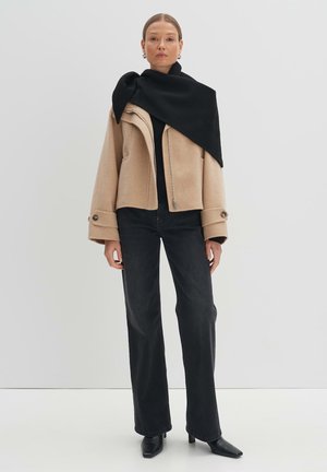 Giacca in misto lana beige con maniche ampie, sciarpa nera appoggiata su un top nero, abbinata a jeans neri a vita alta e stivali neri con tacco.