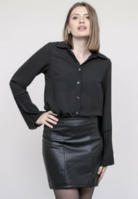 Blouse noire avec fermeture à boutons et longues manches plissées, associée à une jupe droite en simili cuir noir. Tissu lisse avec une finition élégante.