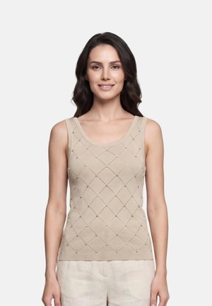 Donna con lunghi capelli scuri che indossa un top beige senza maniche con un motivo a diamante di piccoli borchie e pantaloni chiari, sorridente.