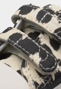 Κοντινό πλάνο ενός σανδαλιού με ασπρόμαυρο μοτίβο, διπλούς ρυθμιζόμενους ιμάντες και λεπτομέρειες ραφής σε λευκό χρώμα.