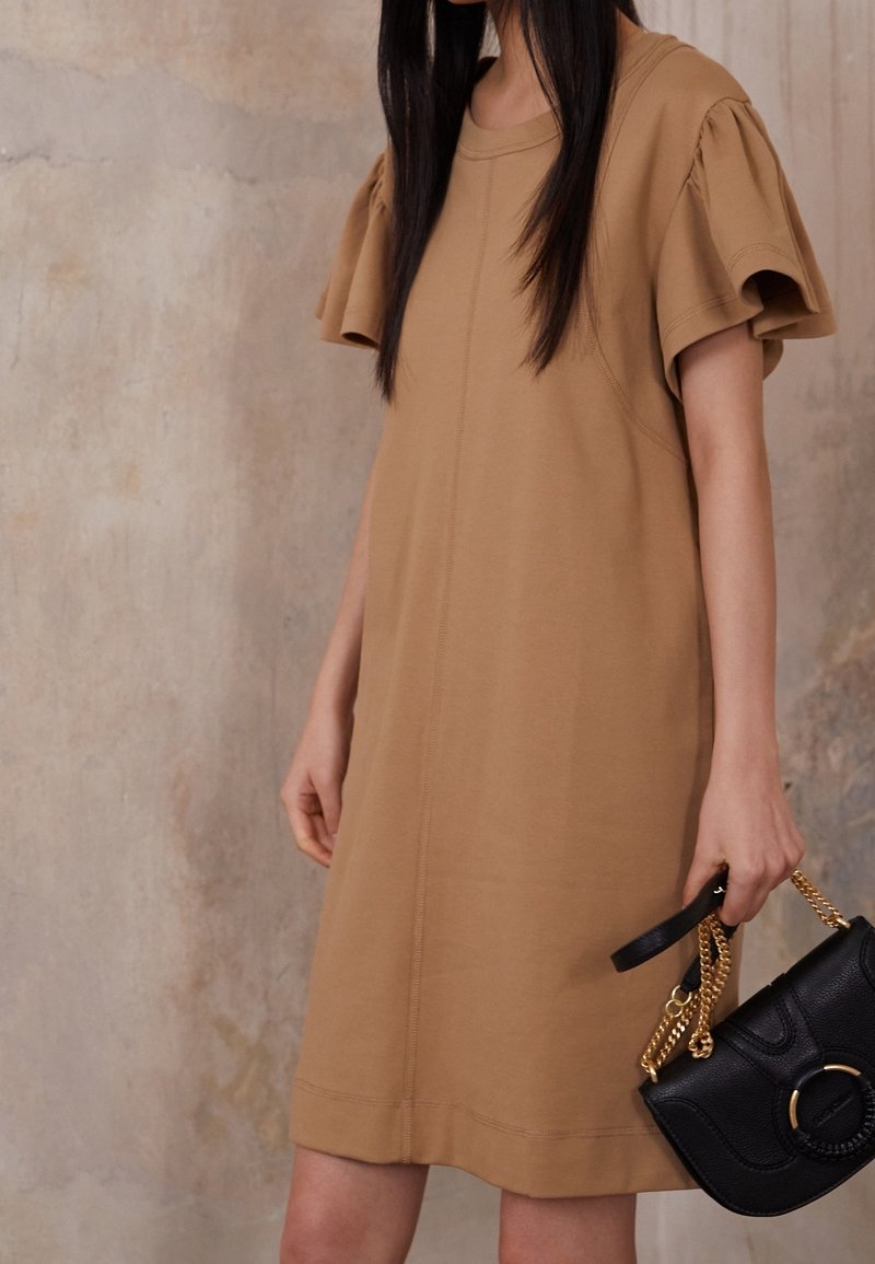 Robe en coton tan avec des manches courtes bouffantes, col ras du cou et détails de couture visibles. Sac en cuir noir à porter à la main avec une chaîne dorée.