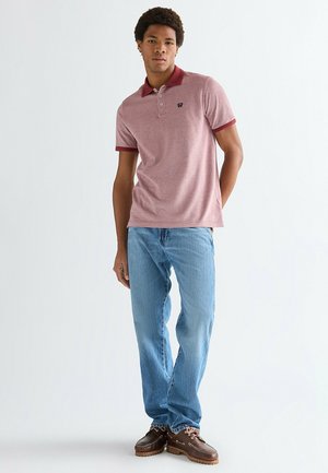 Giovane uomo in piedi che indossa una polo rossa, jeans azzurri e scarpe da barca in pelle marrone su uno sfondo chiaro e uniforme.