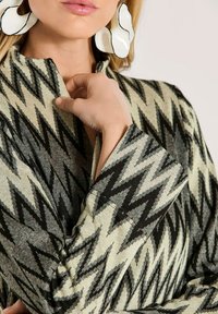 Eine Frau trägt eine Jacke mit schwarz-weißer Zickzackmusterung und große abstrakte weiße Ohrringe. Sie passt mit ihrer Hand den Kragen der Jacke an.