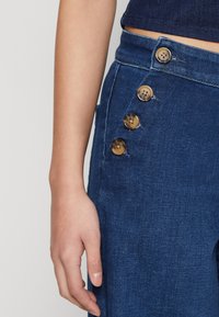 Pantalon en denim bleu foncé avec cinq boutons en laiton sur le côté, présentant une texture lisse et une coupe ajustée, illustré avec une main posée dessus.