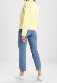 Camicia lunga gialla chiaro a maniche lunghe, vestibilità comoda, abbinata a jeans dritti blu chiaro e sneakers bianche, mostrata da dietro.