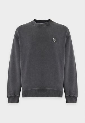Ung mand iført en mørkegrå sweatshirt og blå jeans, der holder en sort mulepose i venstre hånd mod en ensfarvet hvid baggrund.
