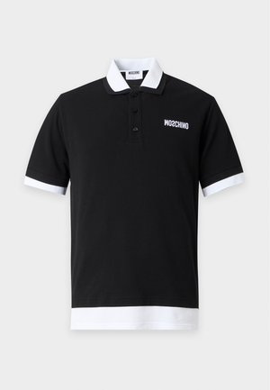 Homme debout portant un polo noir avec col et poignets blancs, un jean bleu et des baskets blanches sur un fond blanc uni.