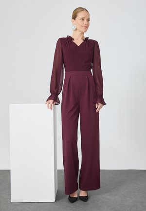 Burgunderfarvet jumpsuit med gennemsigtige lange ærmer, volangmanchetter og V-hals, der har en tætsiddende overdel og brede bukser. Sorte spidse hæle.