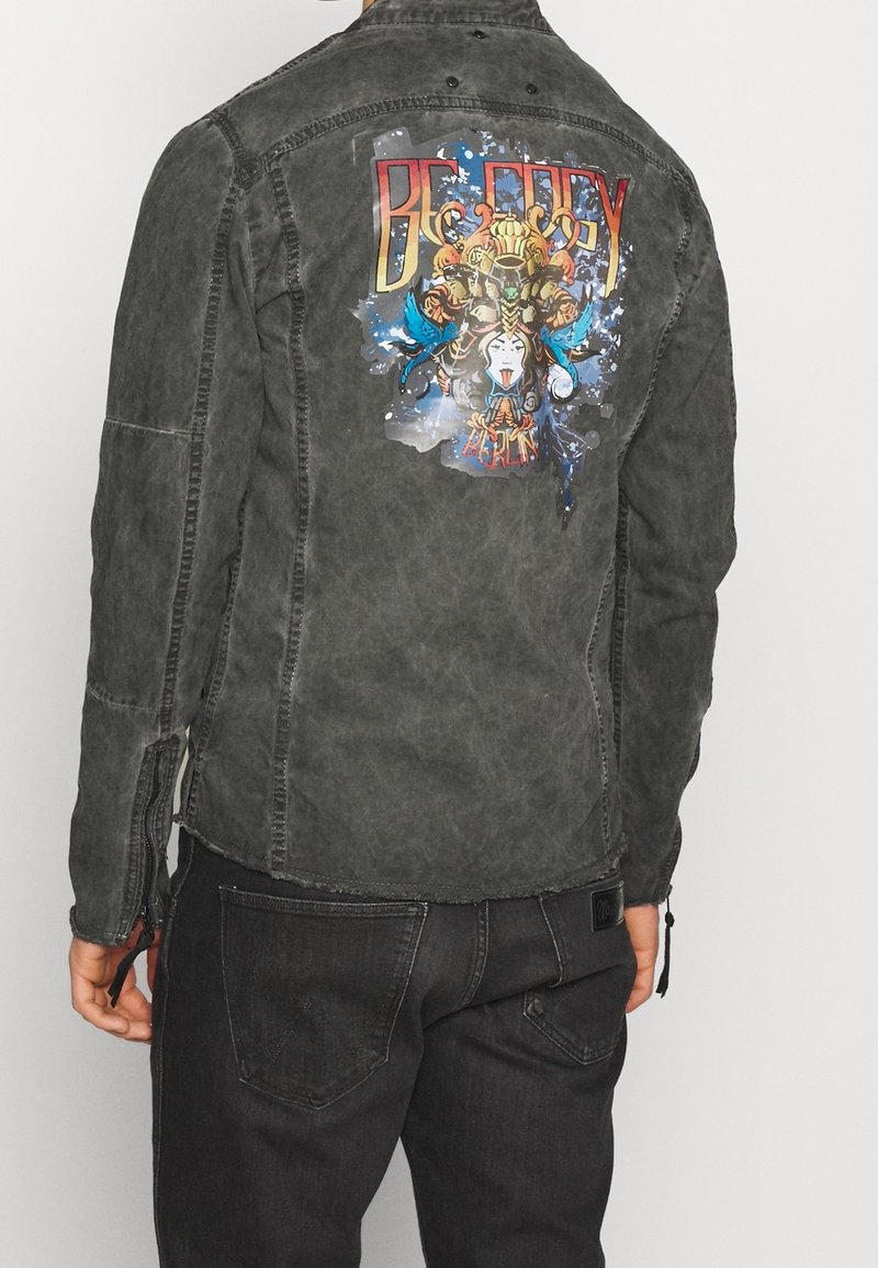 Man som bär en urtvättad svart jacka med färggrafisk konst och texten "BERLIN" på baksidan, tillsammans med mörka jeans.