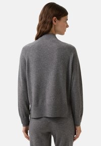 Kvinde iført en grå ribbet turtleneck sweater og matchende bukser, vist fra ryggen mod en ensfarvet baggrund.