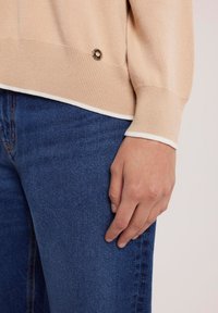 Beżowy sweter z dzianiny z białymi wykończeniami u dolnej krawędzi i mankietów, z okrągłym, metalicznym akcentem w dolnej części. W komplecie z niebieskimi dżinsami.