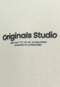 Krémová tkanina s texturovaným úpletem. Černý tištěný text zní "Originals Studio" s dalšími detaily níže v menším písmu.