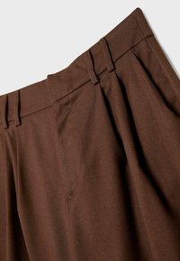 Pantaloni marroni su misura con un design a pieghe, dotati di una cintura con passanti per cintura e una texture liscia. Sono visibili dettagli cuciti vicino alle tasche.