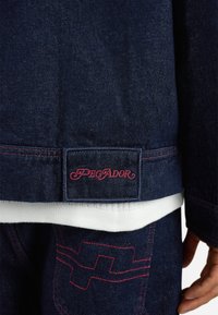 Donkerdenim jacket met roze stiksels, met een "Pegador" label aan de onderkant en een wit onderhemd dat zichtbaar is aan de onderkant.