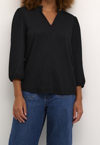 Blouse noire texturée avec un col en V et des manches trois-quarts resserrées, associée à un jean large bleu clair.