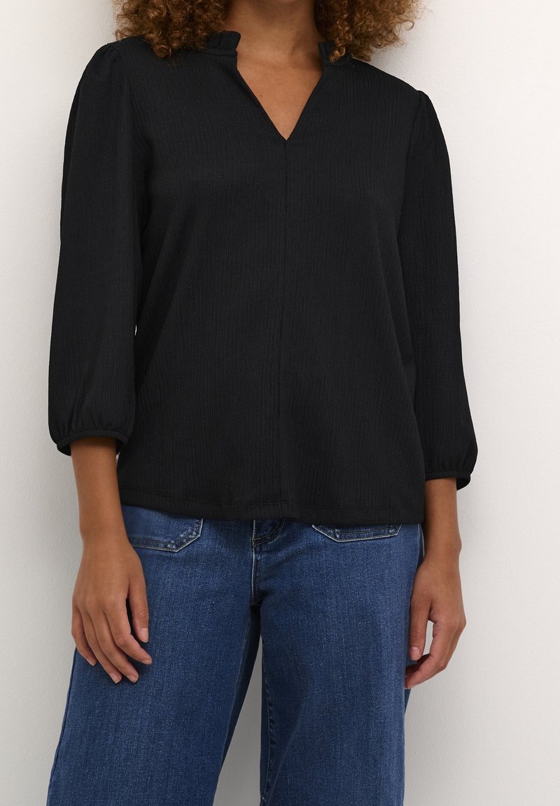 Blouse noire texturée avec un col en V et des manches trois-quarts resserrées, associée à un jean large bleu clair.