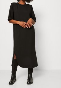 Schwarzes knielanges Kleid mit kurzen Ärmeln, runder Ausschnitt und Seitenschlitz. Aus glattem Stoff gefertigt, getragen mit kniehohen schwarzen Stiefeln.