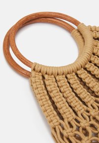 Sac en macramé avec corde en coton beige, doté de poignées circulaires en bois. La texture est tissée avec un motif de nœuds et de franges.