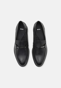 Mocassins en cuir noir avec une finition lisse, dotés d'une barre d'accent en argent, d'un bout arrondi et d'une semelle intérieure rembourrée. Nom de la marque "BOSS" visible à l'intérieur.
