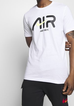 Homme portant un T-shirt blanc avec l'inscription "AIR" et le logo Swoosh de Nike au-dessus du texte "JUST DO IT.", associé à un pantalon noir avec des logos Nike rouges et orange.
