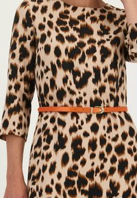 Beige jurk met luipaardprint, voorzien van zwarte en bruine vlekken. Vergezeld van een aansluitende oranje riem met een gouden gesp. Lange mouwen.
