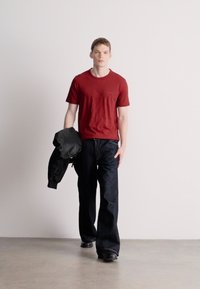 Κόκκινο κοντομάνικο μπλουζάκι, σκούρα μπλε τζιν παντελόνι με φαρδιά πόδια και μαύρο μπουφάν bomber κρατημένο στο χέρι. Λεπτό ύφασμα, απλός σχεδιασμός, casual εμφάνιση.