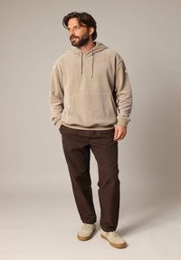 Beige hoodie met een voorzak en een trekkoord. Donkerbruine broek en lichtbeige schoenen met een gestructureerde afwerking.