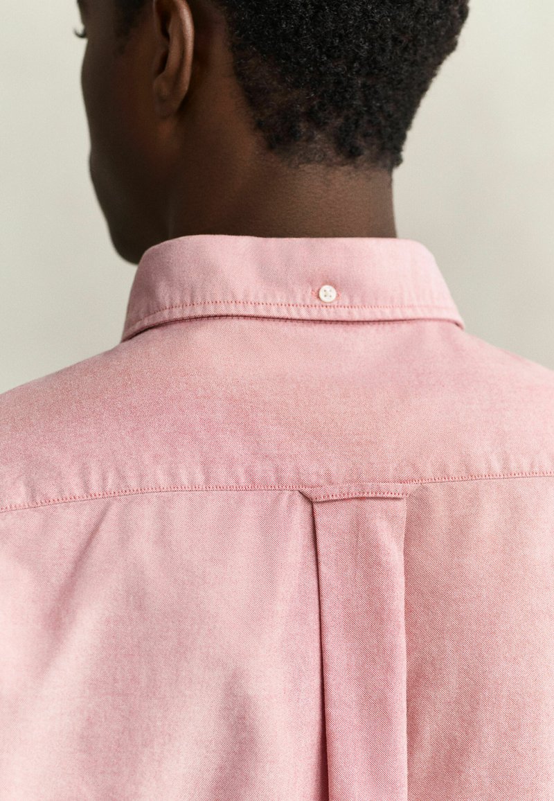 Primer plano de la espalda de una persona que lleva una camisa rosa claro abotonada con un botón en el cuello de la camisa.