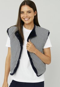 Gilet à carreaux bleu marine et blanc avec garniture en fausse fourrure, ouvert sur le devant et sans manches, porté sur une chemise blanche. Présente des fermetures à pression.