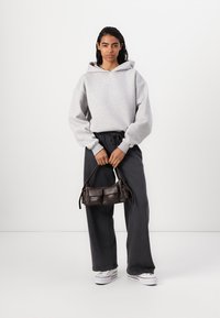 Felpa grigia oversize, pantaloni larghi scuri e sneakers bianche; tiene in mano una piccola borsa scura con due tasche frontali. Texture del tessuto morbida.