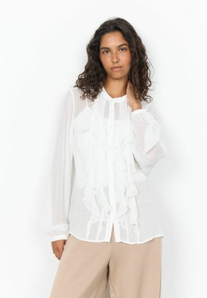Frau mit lockigem dunklem Haar, die eine weiße langärmelige Bluse mit Rüschen und beige Hosen trägt und mit einer Hand ihren Hals berührt.