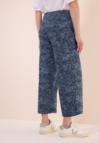 Marinblå vida byxor med ett vitt abstrakt vågmönster, tillverkade av mjukt tyg, kombinerat med vita sneakers med en grön accent.