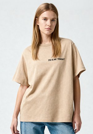 Junge Frau mit glatten blonden Haaren, die ein beiges oversized T-Shirt mit dem Text „YOU & ME TONIGHT“ und blaue Jeans vor einfarbigem Hintergrund trägt.