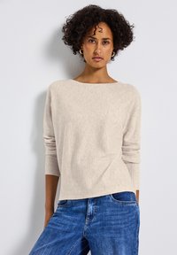 Beige Strickpullover mit weitem Rundhalsausschnitt und gerippten Bündchen, kombiniert mit blauen Jeans, die einen lockeren Sitz und eine weiche Textur haben.