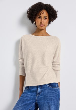 Beige gebreide trui met een brede ronde hals en ribgebreide manchetten, gecombineerd met blauwe jeans, die een relaxte pasvorm en zachte textuur heeft.