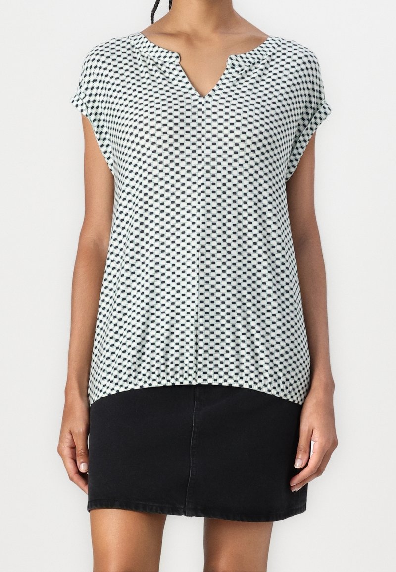 Femme portant un chemisier à manches courtes blanc et noir à motifs et une jupe noire unie sur fond blanc.